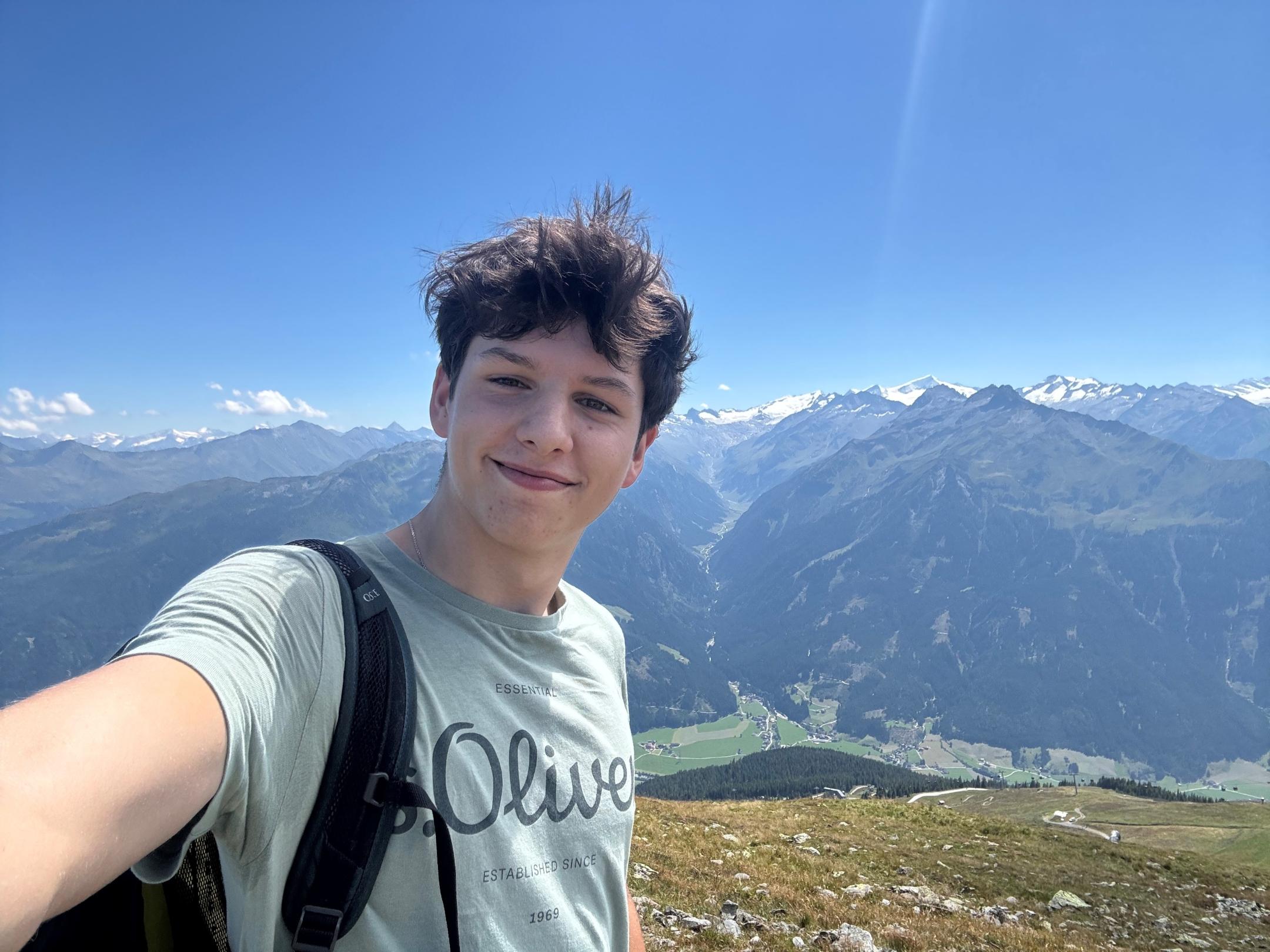 Selfie von einem jungen lächelnden Mann, im Hintergrund eine Gebirgslandschaft
