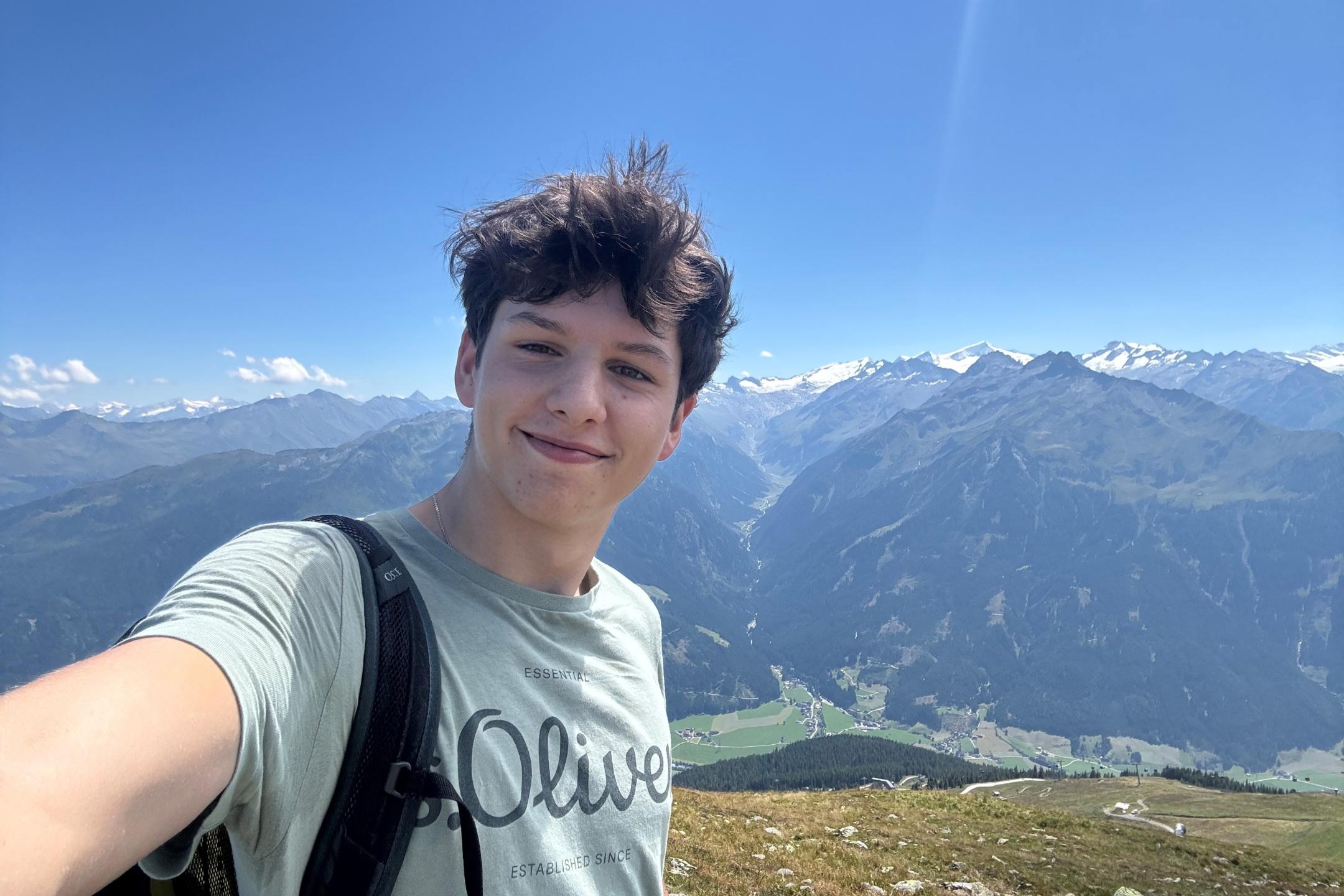 Selfie von einem jungen lächelnden Mann, im Hintergrund eine Gebirgslandschaft