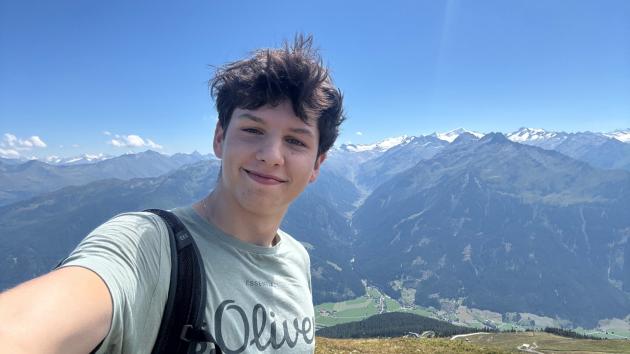 Selfie von einem jungen lächelnden Mann, im Hintergrund eine Gebirgslandschaft