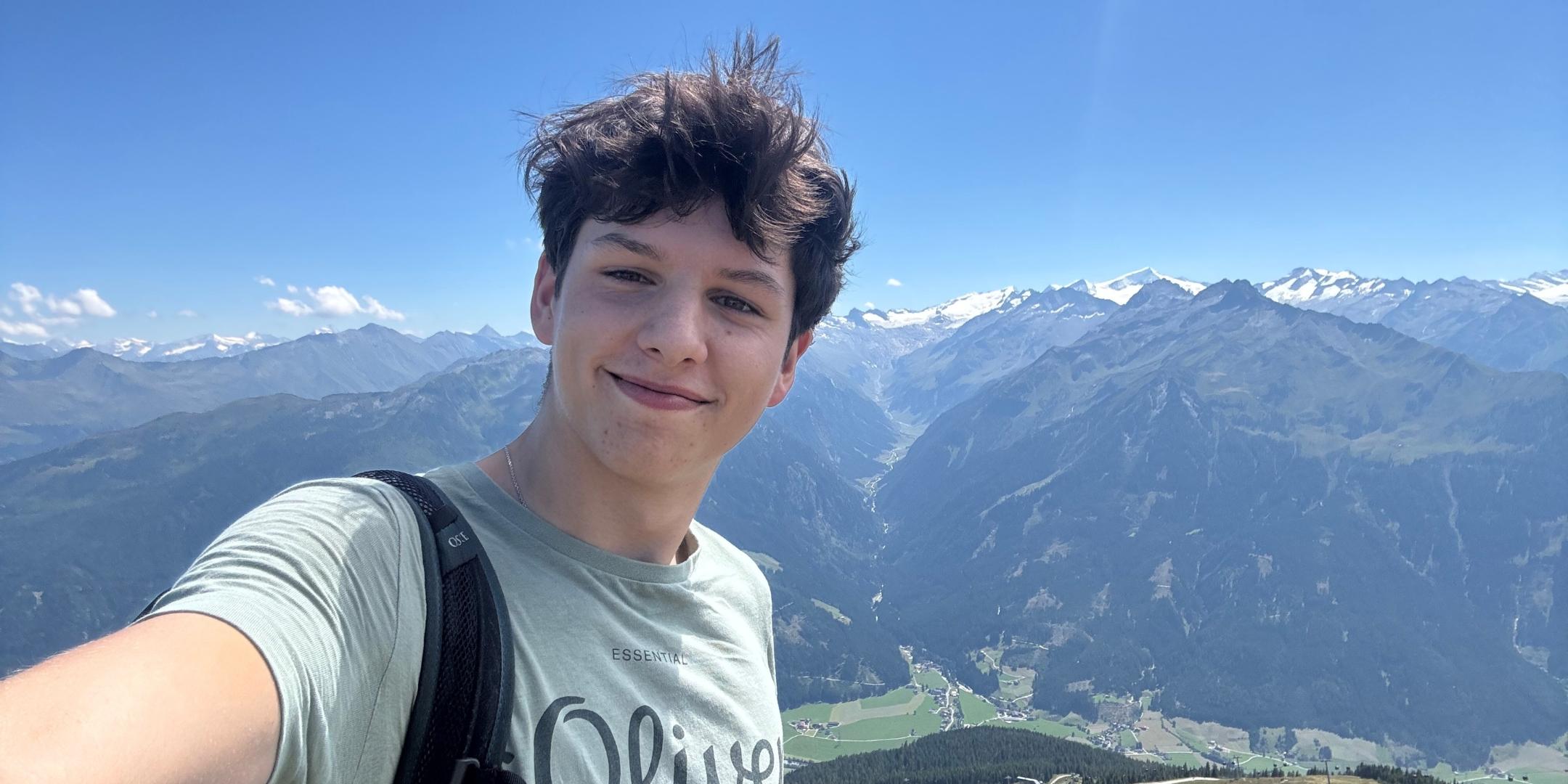 Selfie von einem jungen lächelnden Mann, im Hintergrund eine Gebirgslandschaft